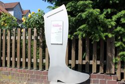 Übergroßer Schusterstiefel mit Beschreibungstafel vor einem Zaun in Wittingen.