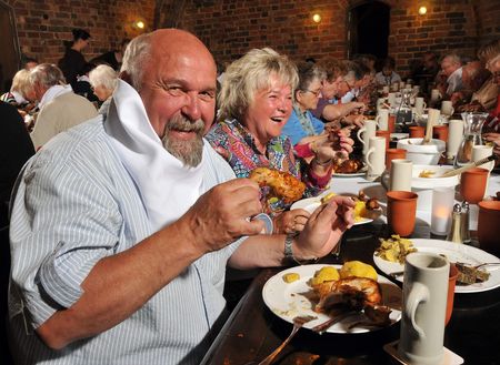 Im Vordergrund einer langen Tafel sitzten ein Mann und eine Frau und essen ein Hähnchenschenkel.