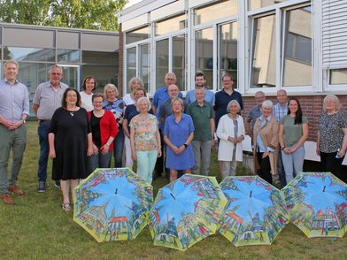Personengruppe vor einem Haus mit aufgespannten Regenschirmen auf dem Boden.