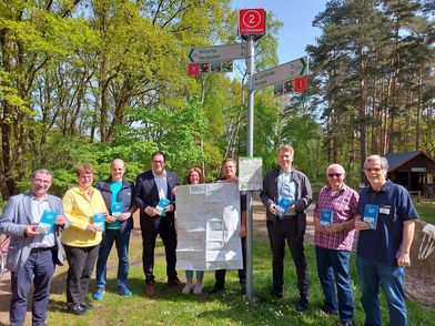 Personengruppe mit einer aufgeklappten Radkarte vor einem Fahrradwegweiser mit Knotenpunkt in der Natur.