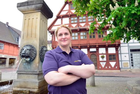 Frau mit verschränkten Armen vor einem Brunnen und einem Fachwerkhaus.